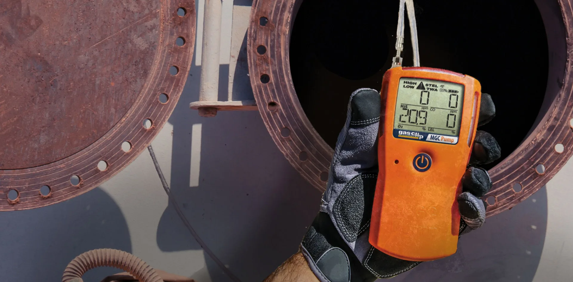 worker wearing safety gloves holds a Gas Clip MGC Pump portable gas detector in front of a rust-colored industrial vessel opening, monitoring air quality for H₂S and oxygen levels during confined space entry.